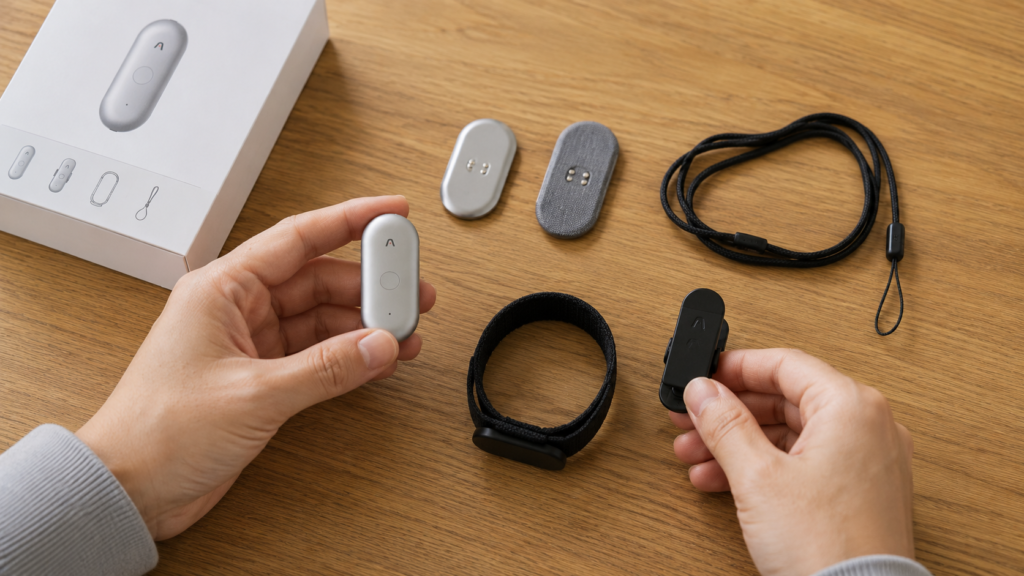 A realistic close-up of the PLAUD NotePin S with its included accessories, including a magnetic pin, clip, wristband, and neck lanyard, arranged on a wooden desk with hands holding the device.