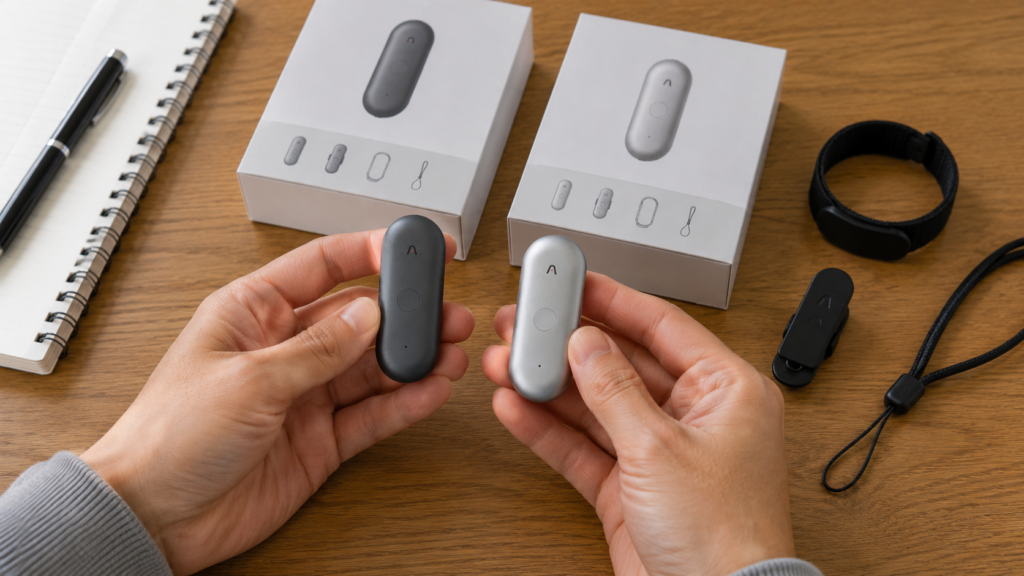 A realistic side-by-side comparison of the PLAUD NotePin and NotePin S held in both hands on a wooden desk, showing their similar size along with included accessories for everyday use.