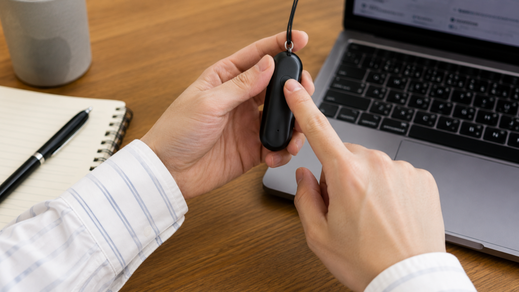 A realistic close-up of a person pressing the physical button on a PLAUD NotePin S wearable voice recorder while holding it naturally beside a laptop and notebook on a desk, demonstrating easy one-touch recording control.