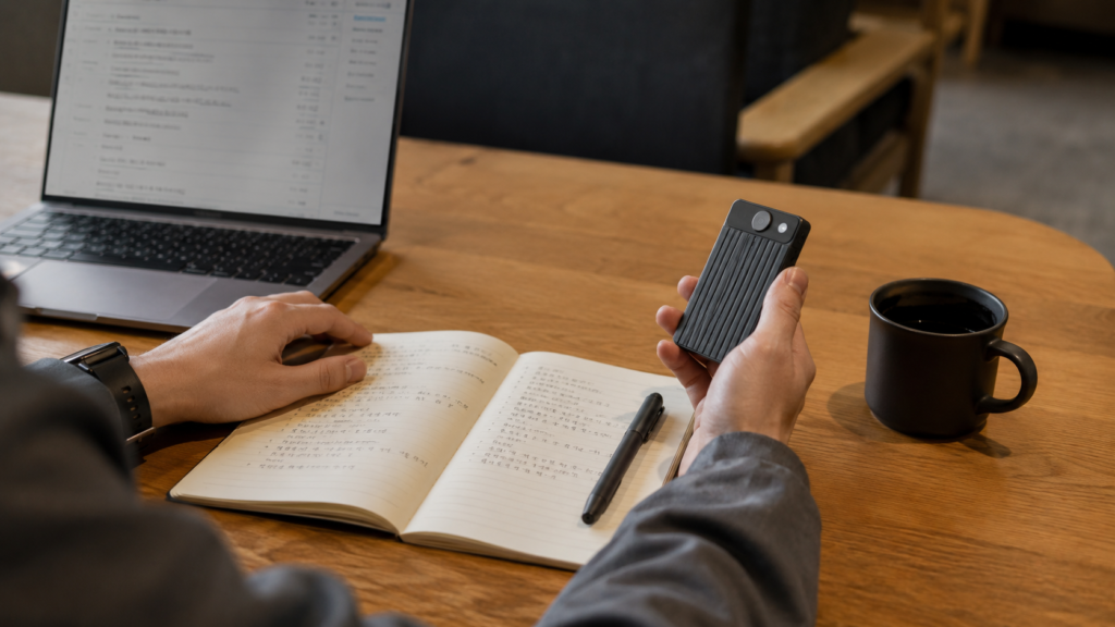 A professional holding a compact AI voice recorder while reviewing meeting notes beside a laptop at a wooden desk.