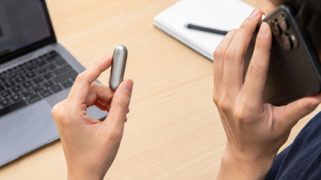 A person holding a PLAUD NotePin wearable AI voice recorder while making a phone call with a smartphone at a desk, illustrating that NotePin is designed for in-person conversations rather than direct phone call recording.